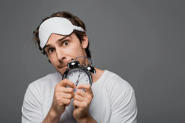 Tensed man in sleep mask holding alarm clock isolated on grey — Stock Photo