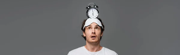 Astonished man in sleep mask looking at alarm clock on head isolated on grey, banner — Stock Photo