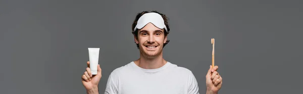 Hombre sonriente con máscara para dormir sosteniendo cepillo de dientes y pasta de dientes aislados en gris, estandarte - foto de stock