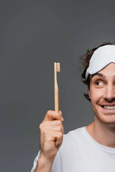 Ausgeschnittene Ansicht eines lächelnden Mannes mit Schlafmaske, der auf die Zahnbürste schaut, isoliert auf Grau — Stockfoto