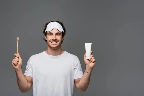 Fröhlicher junger Mann mit Schlafmaske hält Zahnbürste und Zahnpasta isoliert auf grau — Stockfoto