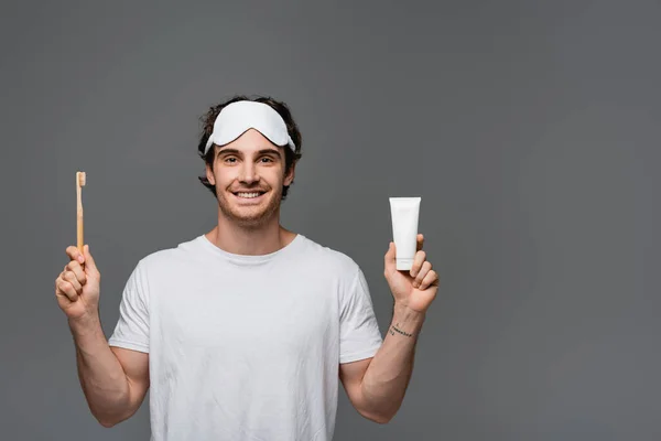 Joyeux jeune homme au masque de sommeil tenant brosse à dents et dentifrice isolés sur gris — Photo de stock