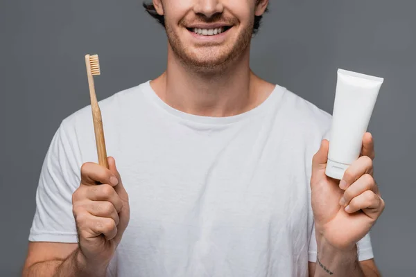 Cropped view of smiling man in white t-shirt holding toothbrush and toothpaste isolated on grey — Stock Photo