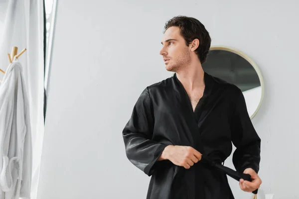 Young man holding belt from satin robe in bathroom — Stock Photo