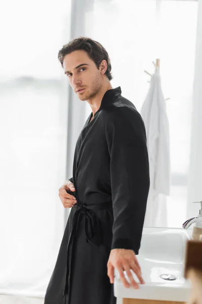 Young man in black sink robe looking at camera near sink in bathroom — Stock Photo