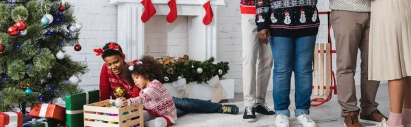 Afrikanisch-amerikanische Kinder spielen mit Christbaumkugeln in der Nähe von Weihnachtsbaum und Familie im Wohnzimmer, Banner — Stockfoto