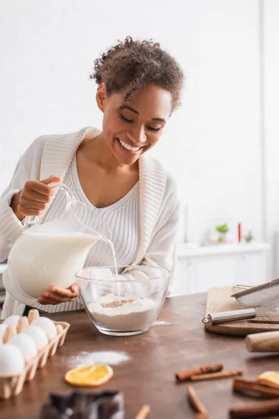 Sorrindo mulher afro-americana derramando leite na farinha perto de paus de canela na cozinha — Fotografia de Stock