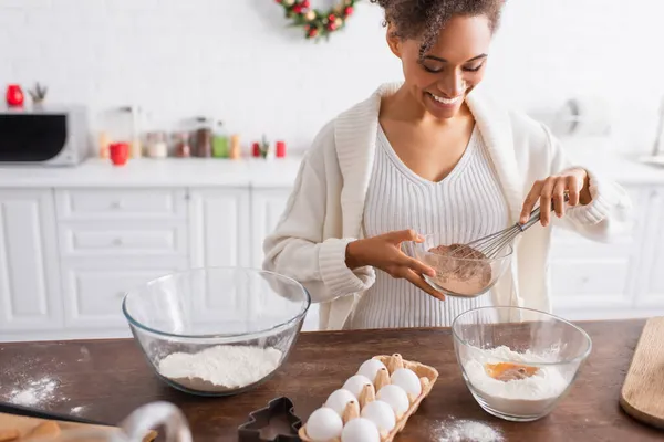 Улыбающаяся африканская американка держит какао во время приготовления пищи рядом с ингредиентами и резаком печенья на кухне — стоковое фото