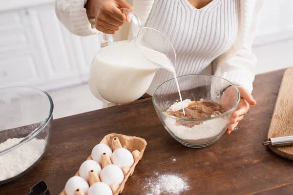 Ausgeschnittene Ansicht einer afrikanisch-amerikanischen Frau, die Milch in Mehl gießt, in der Nähe von Eiern und Ausstecher in der Küche — Stockfoto
