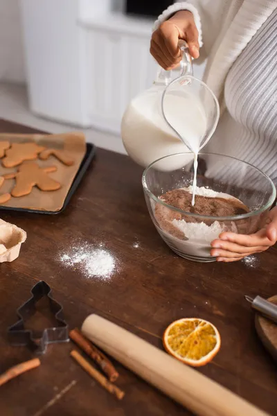 Vue recadrée de la femme afro-américaine versant du lait dans la farine près des biscuits et des bâtonnets de cannelle — Photo de stock