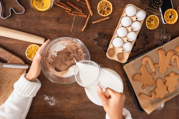 Draufsicht einer afrikanisch-amerikanischen Frau, die Milch in Mehl mit Kakao in der Nähe von Zimtstangen und Weihnachtsplätzchen gießt — Stockfoto