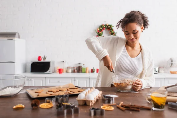 Mujer afroamericana sonriente cocinando cerca de ingredientes y galletas de Navidad en la cocina - foto de stock