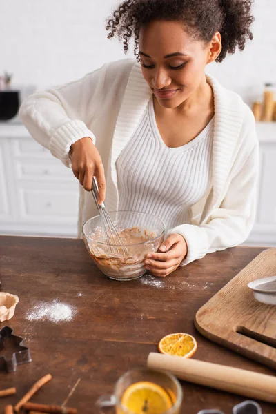 Mujer afroamericana cocinando cerca de palitos de canela borrosa y cortador de galletas en la cocina - foto de stock