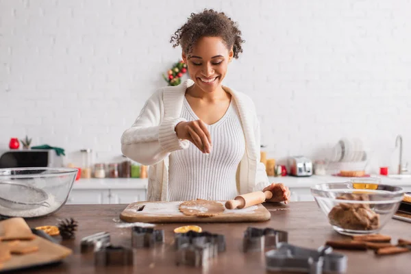 Happy african american woman pouring flour on dough near cookie cutters and cinnamon sticks in kitchen — Stock Photo