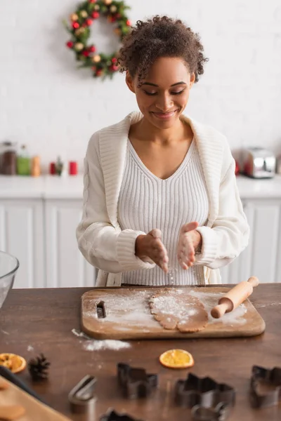 Sourire femme afro-américaine verser de la farine sur la pâte près de rouleau à pâtisserie et coupe-biscuits dans la cuisine — Photo de stock