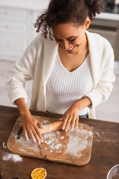 Sorridente donna afroamericana rotolamento pasta sul tagliere vicino fetta di arancia secca in cucina — Foto stock