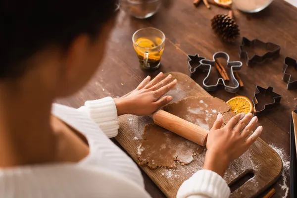 Vue grand angle de la femme afro-américaine floue rouler la pâte près des emporte-pièces de Noël et le thé dans la cuisine — Photo de stock