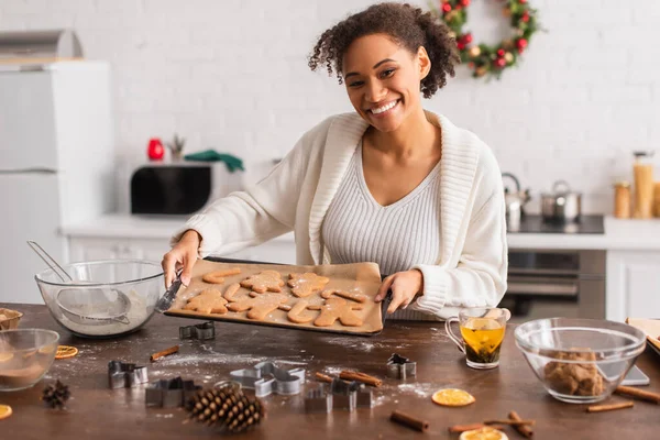 Cheerful african american woman holding christmas cookies on tray near ingredients in kitchen — Stock Photo