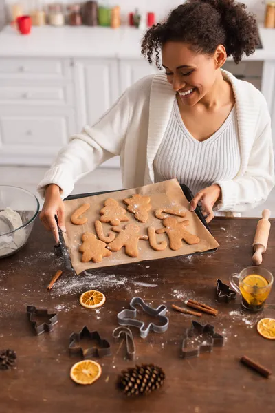 Lächelnde Afroamerikanerin hält Weihnachtsplätzchen auf Tablett neben Zimtstangen und Tee in der Küche — Stockfoto
