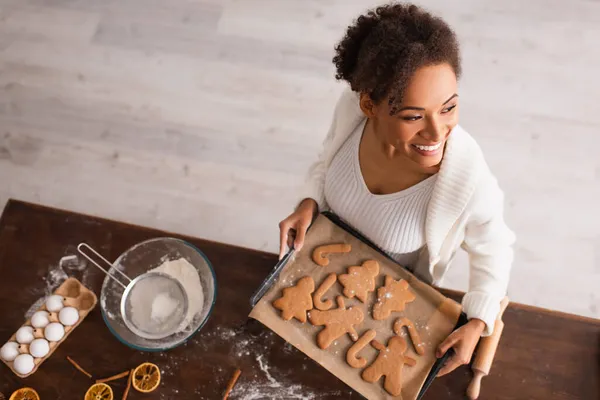 Vue aérienne d'une femme afro-américaine souriante tenant un plateau avec des biscuits de Noël près des ingrédients dans la cuisine — Photo de stock