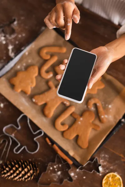 Vista superior de la mujer afroamericana con harina en las manos sosteniendo el teléfono inteligente cerca de las galletas borrosas de Navidad en la cocina - foto de stock