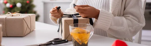 Cropped view of african american woman tying bow on gift near tea and scissors at home, banner — Stock Photo