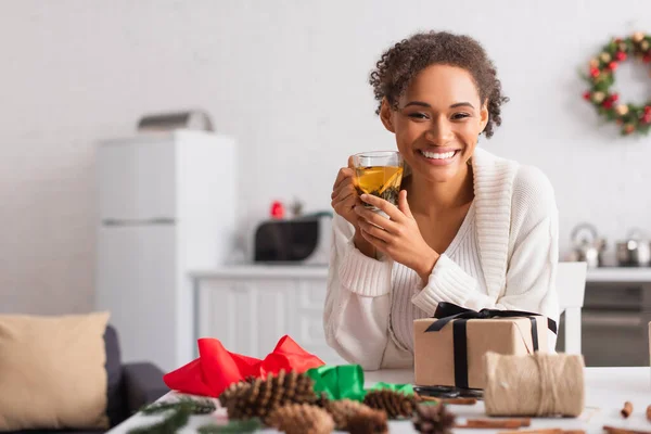 Smiling african american woman holding tea near present and decor at home — Stock Photo