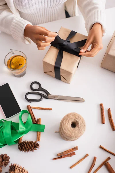 Cropped view of african american woman tying bow on present near decor, smartphone and tea — Stock Photo