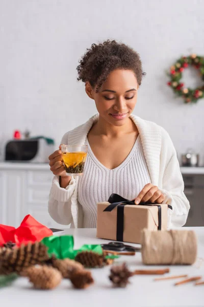 Jovem afro-americana segurando chá enquanto decora o presente de Natal em casa — Fotografia de Stock