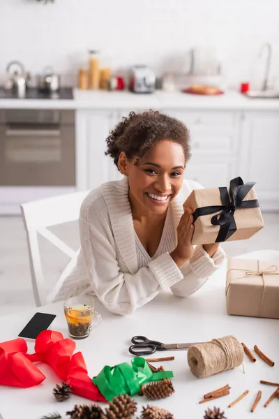 Happy african american woman holding present near decor, tea and smartphone at home — Stock Photo