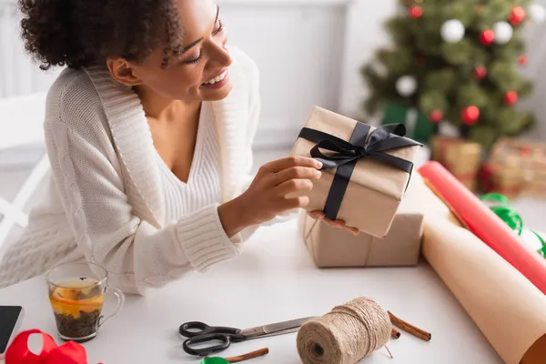 Mujer afroamericana sonriente sosteniendo presente cerca de té, decoración y palitos de canela en casa - foto de stock