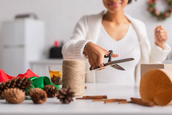 Vista recortada de la borrosa mujer afroamericana cortando el cordel mientras decoraba el regalo de Navidad - foto de stock