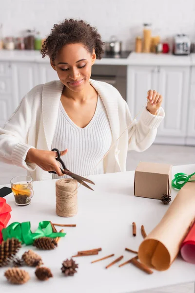 Young african american woman cutting twine while decorating gift near tea and cinnamon sticks — Stock Photo