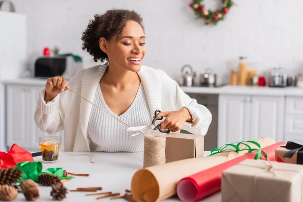 Positive african american woman cutting twine near decor and gifts at home — Stock Photo