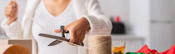 Vista recortada de la borrosa mujer afroamericana cortando el cordel mientras decoraba el regalo de Navidad, pancarta - foto de stock