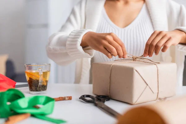 Cropped view of african american woman tying twine on gift near tea and scissors at home — Stock Photo