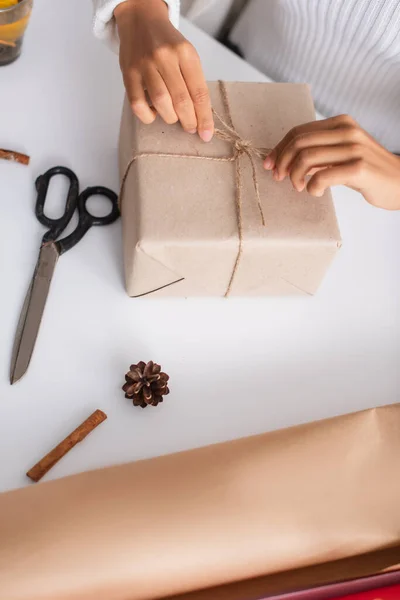 Cropped view of african american woman tying twine on present near pine cone and scissors — Stock Photo