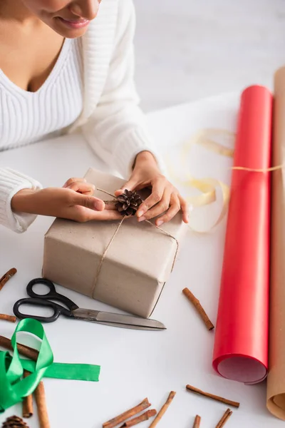 Cropped view of african american woman decorating present with pine cone near scissors at home — Stock Photo