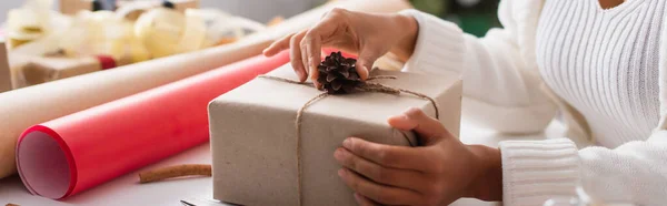 Cropped view of african american woman decorating present with pine cone near wrapping paper, banner — Stock Photo