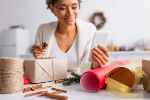Smartphone en la mano de una mujer afroamericana borrosa decorando el regalo de Navidad en casa - foto de stock