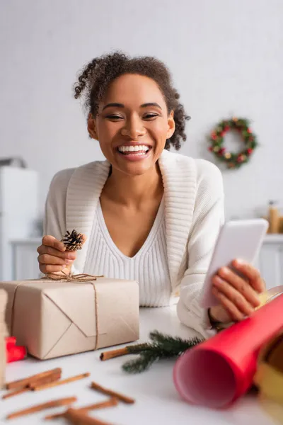 Cheerful african american woman holding smartphone while decorating present at home — Stock Photo