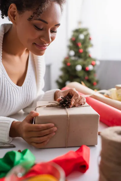 Lächelnde Afroamerikanerin dekoriert Geschenkbox mit Tannenzapfen in der Nähe von Dekor zu Hause — Stockfoto