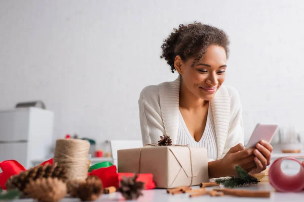 African american woman using smartphone near present and blurred decor at home — Stock Photo
