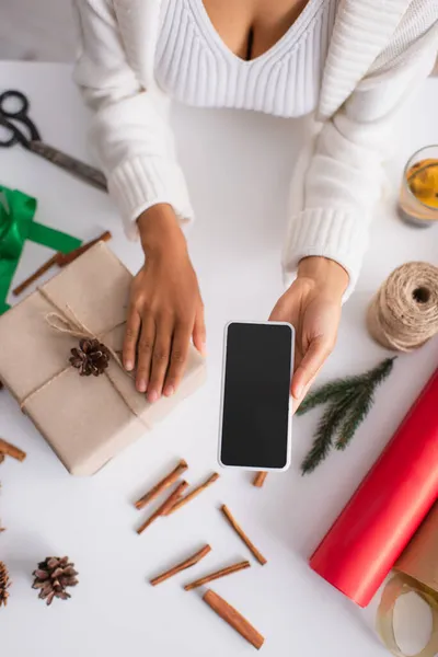 Vista dall'alto della donna afro-americana che tiene smartphone vicino all'arredamento presente e natalizio a casa — Foto stock