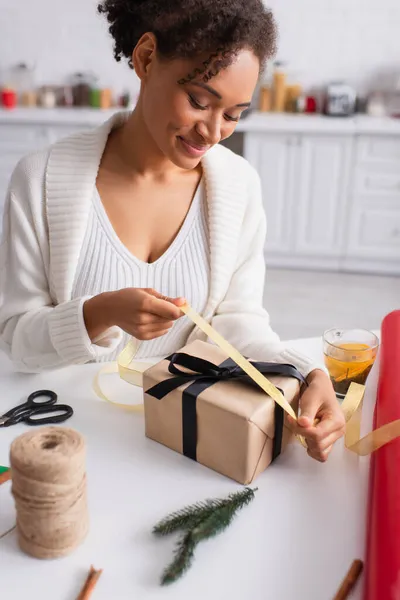 Happy african american woman holding ribbon while decorating christmas gift at home — Stock Photo