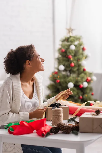 Side view of cheerful african american woman decorating present near blurred christmas tree — Stock Photo