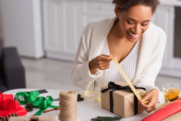 Positive afrikanisch-amerikanische Frau hält Schleife beim Dekorieren von Geschenken in der Nähe einer Tasse Tee zu Hause — Stockfoto