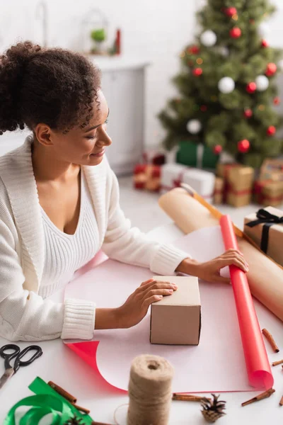 African american woman holding present and wrapping paper near christmas decor at home — Stock Photo