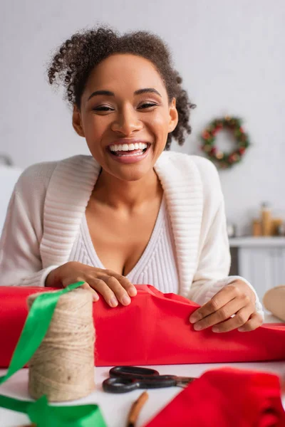 Positive african american woman looking at camera while wrapping christmas gift at home — Stock Photo