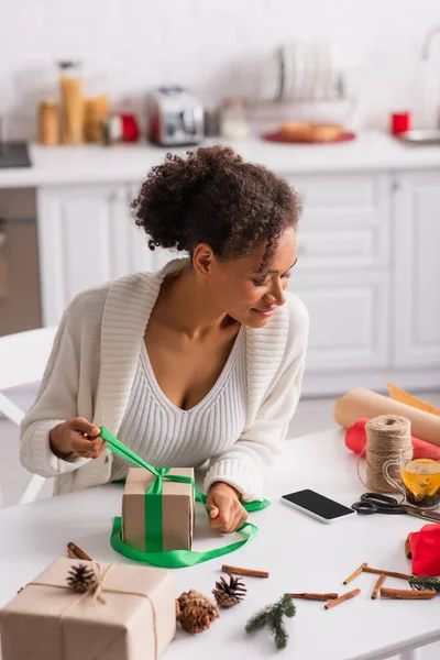 Donna afroamericana che guarda smartphone mentre arreda presente vicino al tè a casa — Foto stock